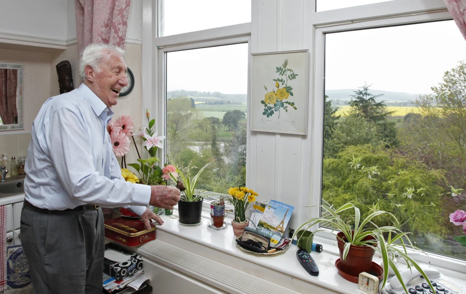A gentleman is stood in the window looking at plants on the sill
